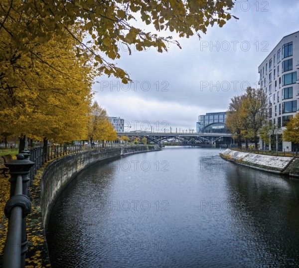 View along a quiet canal, lined with glowing yellow autumn trees and modern architecture, under a cloudy sky, at Berlin Central Station, 10557 Berlin, Germany