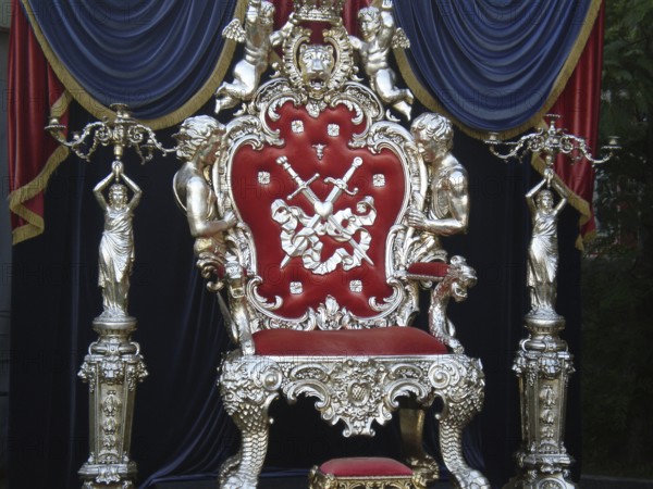 A luxurious royal-style chair with elements such as a crown, lion, angels, and swords, patterns, unique decorations such as statues, and velvet curtains in the background. This vintage installation was located in a public space on the Yalta embankment. Yalta, Crimea.Velvet luxury throne on platform