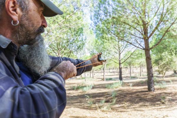 Man with a long beard holding a slingshot, aiming a projectile in a forest environment. Concept of outdoor hunting skill