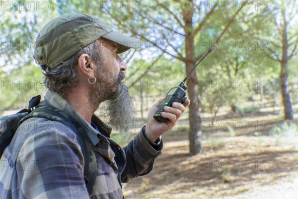 Man with beard and cap holding a walkie-talkie, communicating during an outdoor activity in a natural pine forest