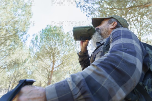 Man with beard and cap drinking water from a green canteen, hydrating during a trekking activity in a pine forest