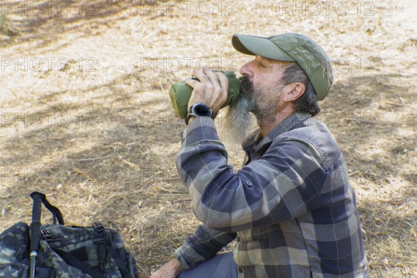 Mature bearded hiker drinking from a canteen during a trekking break in sunlit forest, resting and rehydrating on trail (outdoors)