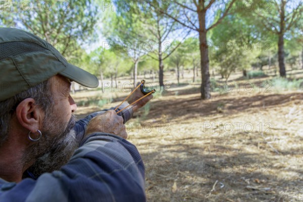 Bearded man in cap aims a slingshot in a sunlit pine forest, focused on target during outdoor bushcraft and hunting practice