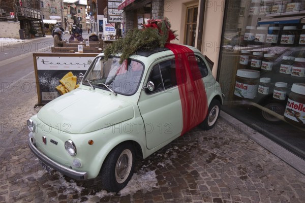 In Livigno in Lombardy in Italy, a small vintage Fiat, decorated as a Christmas present, stands in front of a shop window with huge Nutella glasses