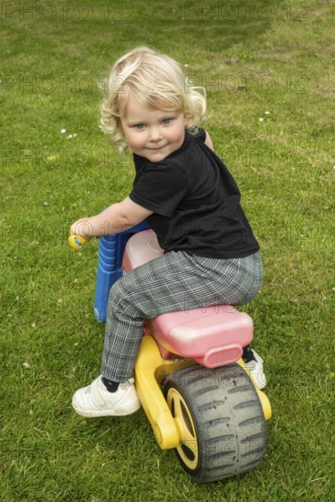 Blond Nordic girl, 3 years old, on a toy vehicle and looking back smiling in Ystad Municipality, Skåne County, Sweden, Scandinavia