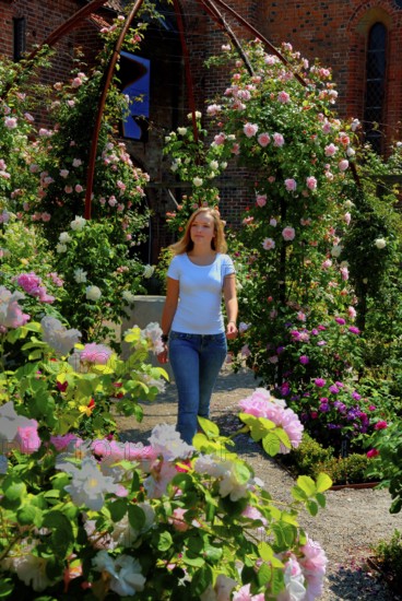 Blonde young beautiful woman walking among roses in the Rosary at the monastery in Ystad, Skåne County, Sweden, Scandinavia
