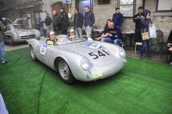 Silver Porsche 550 Spyder with two helmeted drivers drives across the green carpet at the rally, surrounded by an old town crowd, Mille Miglia 2016, time control, checkpoint, SAN MARINO, classic car racing. San Marino, Italia