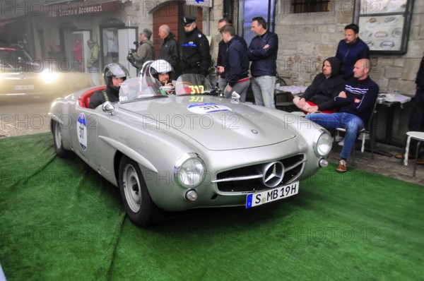 Silver 190 SL Roadster with helmeted occupants slowly across the green carpet, watched by spectators in front of stone facades, Mille Miglia 2016, time control, checkpoint, SAN MARINO, classic car racing. San Marino, Italia