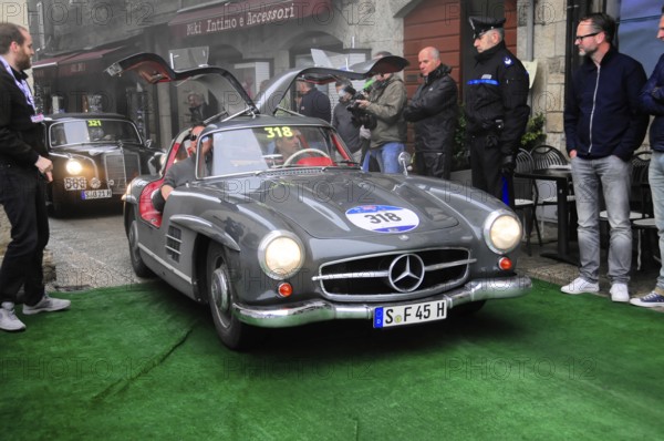 Dark gray 300 SL with open swing doors stops on the green carpet, surrounded by spectators and police in front of historic house facades, Mille Miglia 2016, time control, checkpoint, SAN MARINO, classic car racing. San Marino, Italia