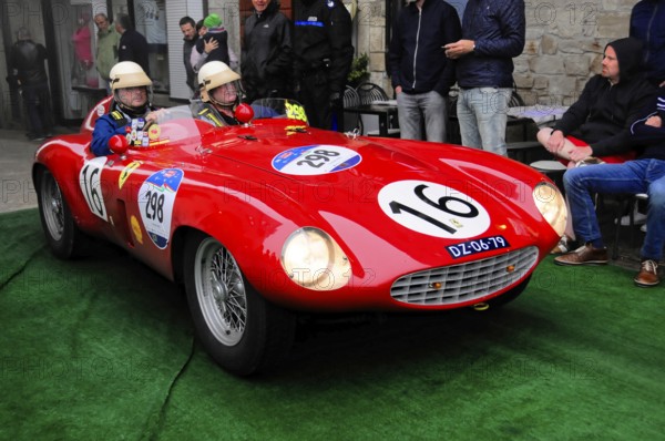 Red racing barchetta with start number 16 glides across the green carpet, two drivers wearing vintage helmets, lively rally atmosphere in the old town, Mille Miglia 2016, time control, checkpoint, SAN MARINO, classic car racing. San Marino, Italia