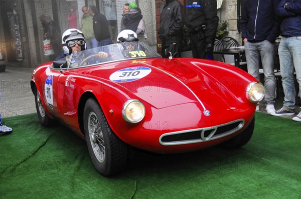 Red roadster with two helmeted drivers rolls across the green carpet at the rally, flanked by spectators in front of gray old town facades, Mille Miglia 2016, time control, checkpoint, SAN MARINO, classic car racing. San Marino, Italia