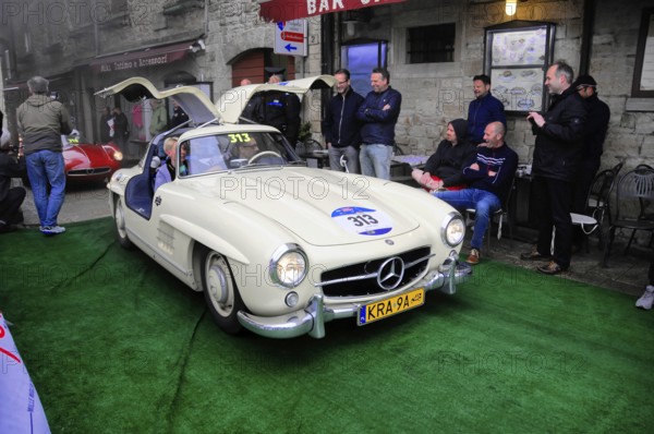 Creamy white 300 SL with open double doors poses on the green carpet, surrounded by smiling spectators in the old town, Mille Miglia 2016, time control, checkpoint, SAN MARINO, classic car racing. San Marino, Italia