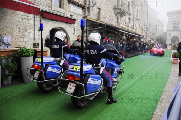 Police motorcycles escort a classic car rally through the stone old town, audience lines the green carpet, Mille Miglia 2016, time control, checkpoint, SAN MARINO, classic car racing. San Marino, Italia