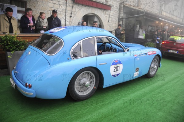 Light blue vintage racing car with start number 201 rolls across green carpet through a historic alley, surrounded by spectators and light fog, Mille Miglia 2016, time control, checkpoint, SAN MARINO, classic car racing. San Marino, Italia
