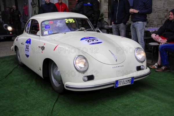 White Porsche 356 with glowing headlights drives across green carpet during a vintage car rally, watching passers-by, Mille Miglia 2016, time control, checkpoint, SAN MARINO, classic car racing. San Marino, Italia
