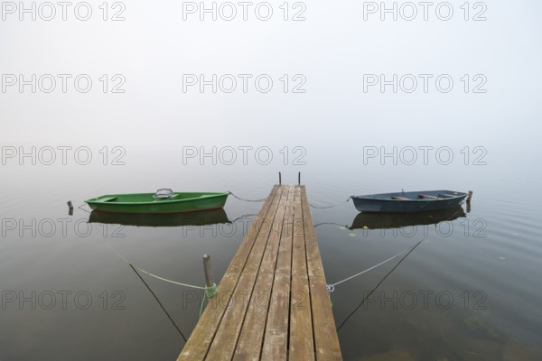 Two boats are moored on a wooden dock in a foggy lake, quiet and peaceful atmosphere, Hopfensee, Ostallgäu, Bavaria, Germany