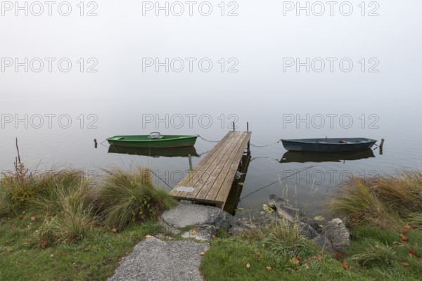 Two boats and a jetty on foggy lakeside with grass and stones, Hopfensee, Ostallgäu, Bavaria, Germany