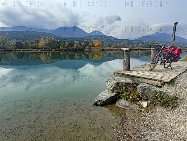 An idyllic lake with a bicycle on a jetty, surrounded by autumnal landscape and mountains, Drau, Villach, Drau cycle path, Carinthia, Austria