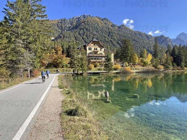 Idyllic autumn scene with a clear lake surrounded by trees and mountains, cyclists on a narrow road, cycle tourism, Lake Toblach, Lake Dobbiaco, South Tyrol, Italy