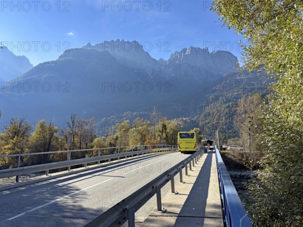 A yellow bus crosses a bridge under a blue sky, surrounded by autumnal mountains, Lienz Dolomitem, Drau, Drauradweg, Lienz, Austria