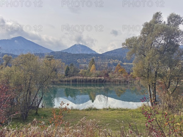 A lake in autumn surroundings, surrounded by mountains and trees under an overcast sky, Drau, Villach, Drauradweg, Carinthia, Austria
