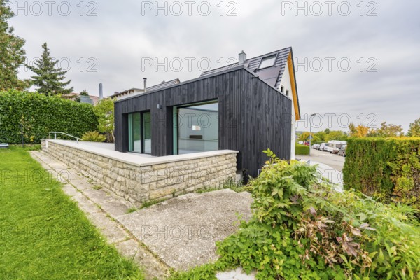 Black, modernly clad extension with large glass areas on lawn and stone wall in a quiet suburban location with cloudy sky, house renovation, air conditioning renovation, Metzingen, Germany