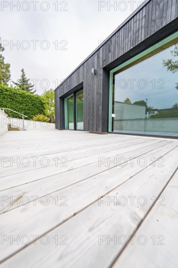 Deep view along the wide wooden terrace of the modern, black-clad building with floor-to-ceiling windows, house renovation, air-conditioning renovation, Metzingen, Germany