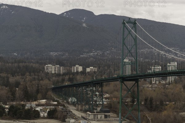 A long suspension bridge connecting a cityscape with mountainous surroundings under an overcast sky in West Vancouver, British Columbia, Canada