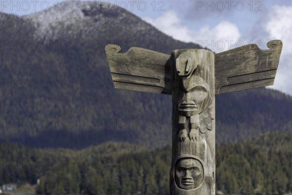 Wooden totem pole stands in front of scenic mountain and forest backdrop under a cloudy sky in Tofino, British Columbia, Canada