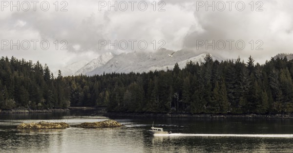 A boat navigates a still waterway lined with forest under a cloudy sky with mountains in the distance in Tofino, British Columbia, Canada