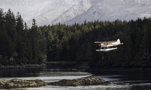 A seaplane flies above rocky shores and dense forest with snow-capped mountains in the background in Tofino, British Columbia, Canada