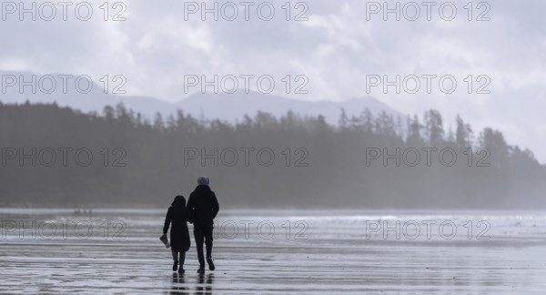 A couple strolls on a misty beach with forest and mountains in the distance in Tofino, British Columbia, Canada