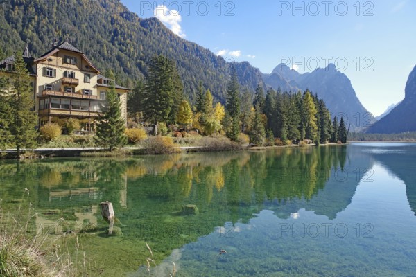 Waterfront villa on a clear mountain lake reflected against rugged peaks, sunny autumn colors glow in the calm water, Lake Toblach, Lake Dobbiaco, South Tyrol, Italy