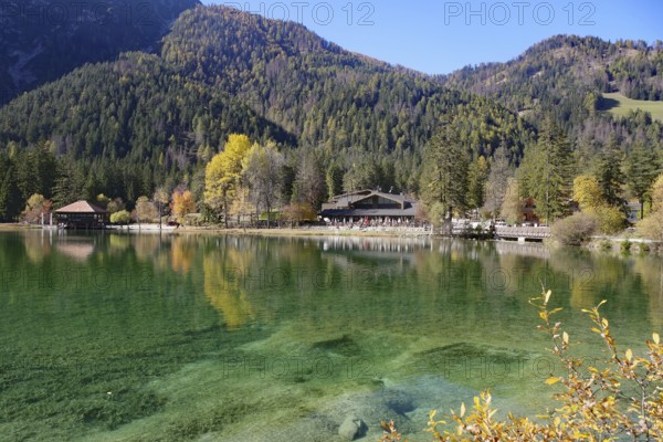 Clear mountain lake with emerald green water, autumnal forest and hut on the shore under blue sky, Lake Toblach, Lake Dobbiaco, South Tyrol, Italy