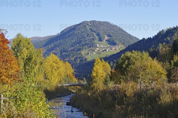 A stream snakes through an autumnal valley, on the sunny mountainside there is a village under deep blue sky, Drava, Drau cycle path, South Tyrol, Italy