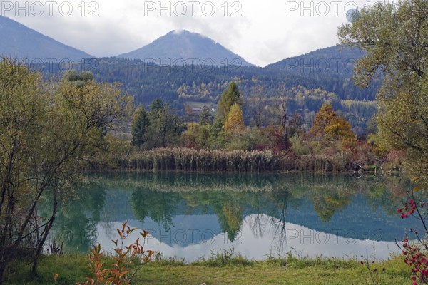 Quiet lake with turquoise-green water, autumnal shore and mountain scenery under cloudy sky, Drau, Drauradweg, Carinthia, near Villach, Austria