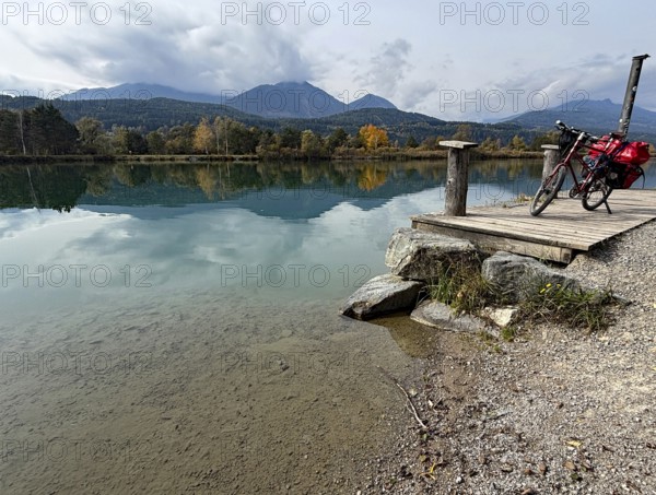 A bicycle is leaning on a footbridge over a still lake, mountains and clouds are reflected in the clear water, Drau, Drau cycle path, Carinthia, near Villach, Austria