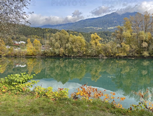 Calm river reflecting autumnal trees and mountains under cloudy sky, Drau, Drauradweg, Carinthia, near Villach, Austria