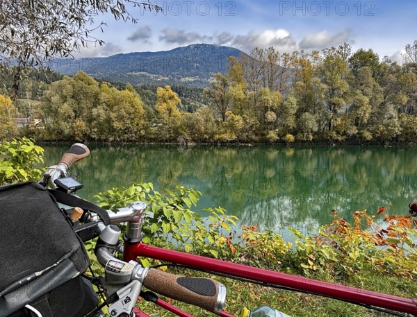 View over the handlebar of a shimmering green river in front of autumn trees and mountains, bicycle tourism, Drau, Drau cycle path, Carinthia, near Villach, Austria