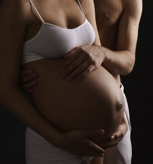 Pregnant young long haired woman staying close to her husband both tenderly looking at her belly and holding their hands on it