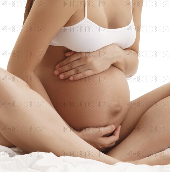 Young long haired pregnant brunette in white top sitting on bed posing holding her hands on her belly studio shot