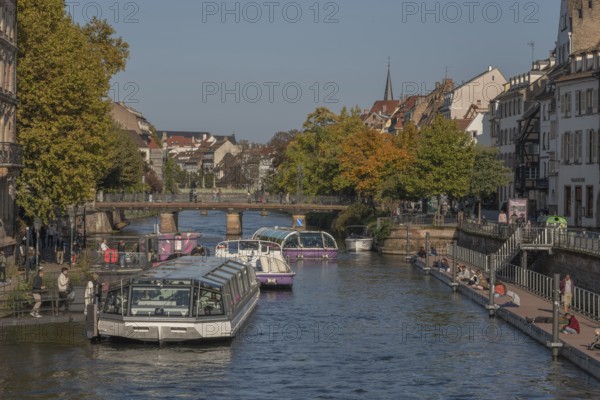 Tourist boat docks on the Ill and transports visitors who enjoy the view of Strasbourg's sights and trees in autumn. The scenery is peaceful and sunny. Bas Rhin, Alsace, France