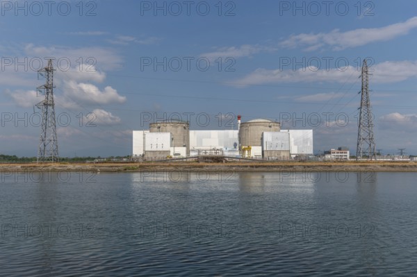 A large industrial nuclear power plant is located on a quiet water front. Two tall cylindrical structures dominate the skyline, with overhanging power lines and distant clouds that ennoble the blue sky. Fessenheim, Haut Rhin, Alsace, France