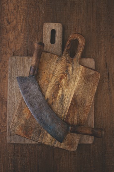 A large curved knife rests on top of two wooden cutting boards on a rustic surface. The knife shows wear, while the boards display unique grain patterns, adding character to the scene