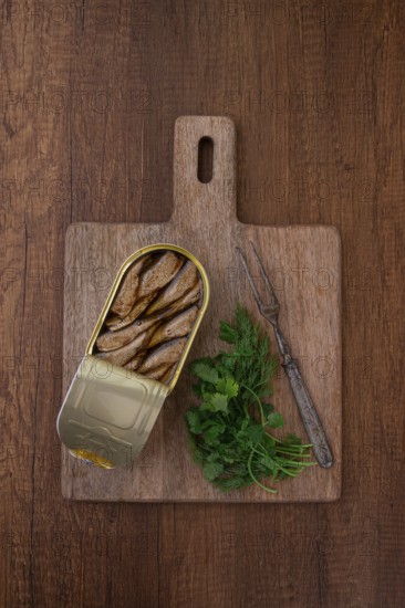 An open can of sprats, appetizer, on a wooden cutting board, with a fork and herbs, top view