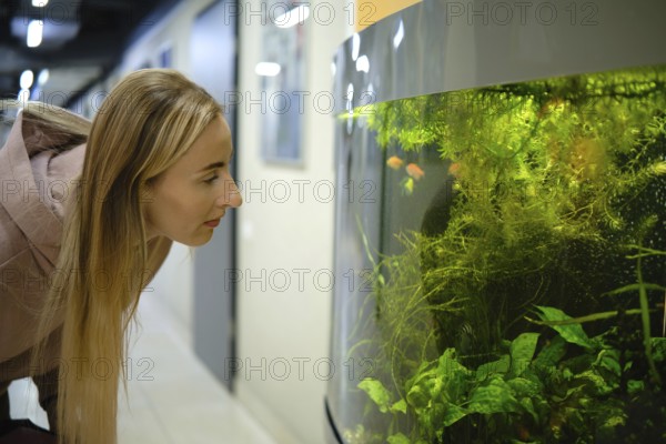 A woman with long hair leans forward, peering into a large aquarium filled with vibrant fish and lush green plants. The setting is a contemporary corridor, creating a calm scene