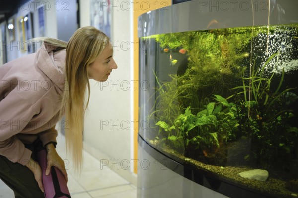 A woman leans forward, observing vibrant fish swimming in a large aquarium. The setting is bright and modern, with green plants enhancing the underwater scene and creating a calm atmosphere