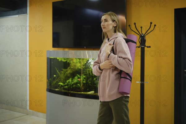 A woman stands in a fitness studio holding a yoga mat. She wears a cozy hoodie and looks ready for her yoga class. An aquarium adds life to the space, creating a serene atmosphere