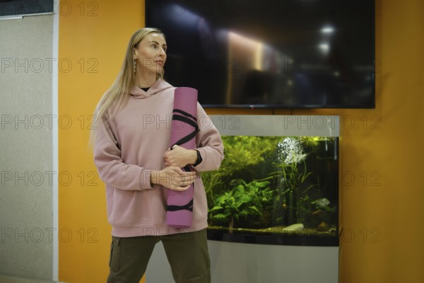 A woman stands in a fitness studio, holding a yoga mat, looking inspired. An aquarium filled with plants and fish is behind her, adding a calming atmosphere to the space during a workout