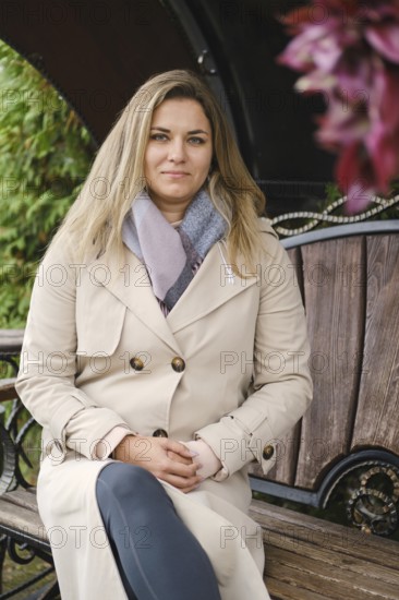 A woman sits calmly on a wooden bench, wearing a beige trench coat and gray scarf. She exudes a relaxed demeanor against a backdrop of greenery and flowers, enjoying a peaceful moment outdoors